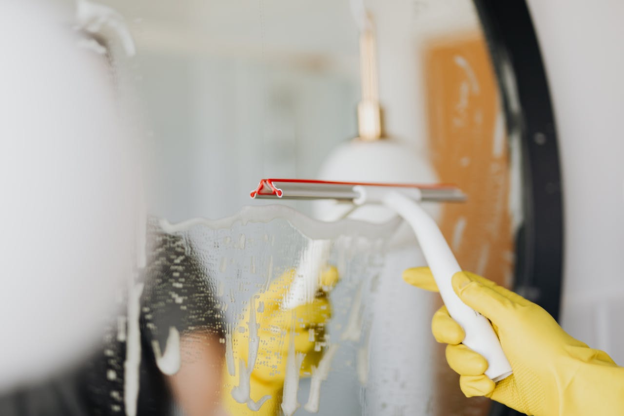 services-05 Anonymous person in yellow rubber gloves wiping water by squeegee from mirror in bathroom